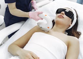 Crop faceless woman in medical uniform and gloves using laser tool on young woman wearing safety glasses and towel lying on couch in hospital